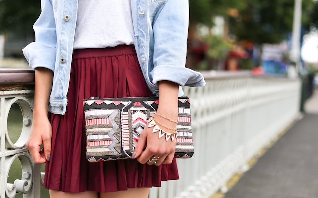 Girl wearing a plum red skirt holding a small bag while walking on a bridge 
