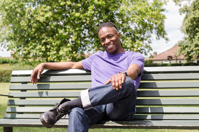Nigel Clarke wearing a purple shirt smiling and sitting on a park bench 