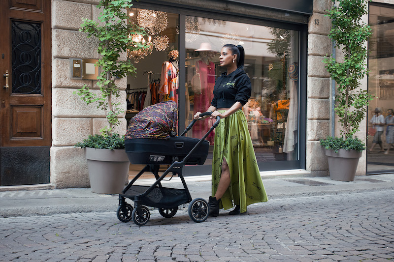 Georgette Polizzi wearing a green skirt holding a NUNA stroller in rainbow colours posing for mums magazine 
