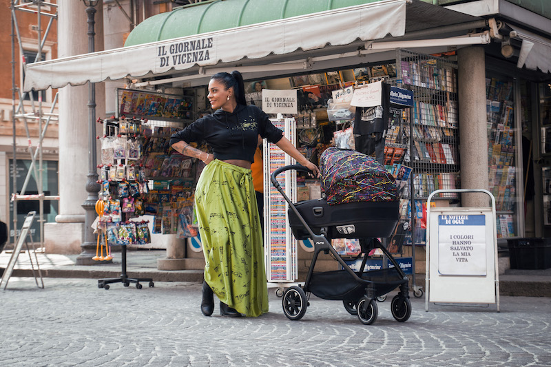 Georgette Polizzi wearing a green skirt holding a NUNA stroller in rainbow colours posing for mums magazine 