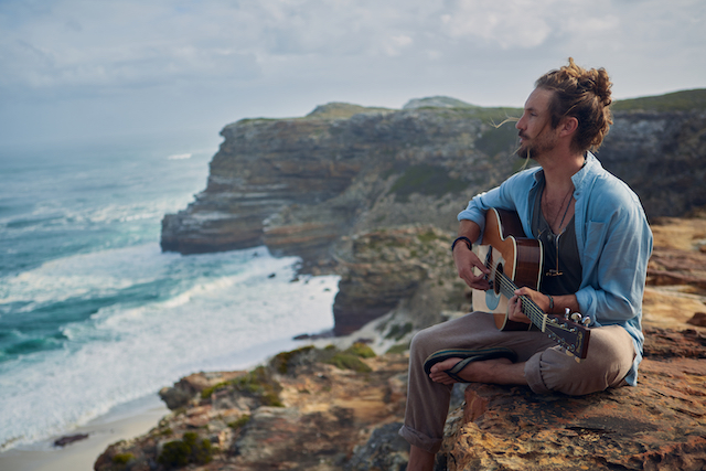 Jeremy Loops wearing a denim shirt and playing the guitar while gazing the sea