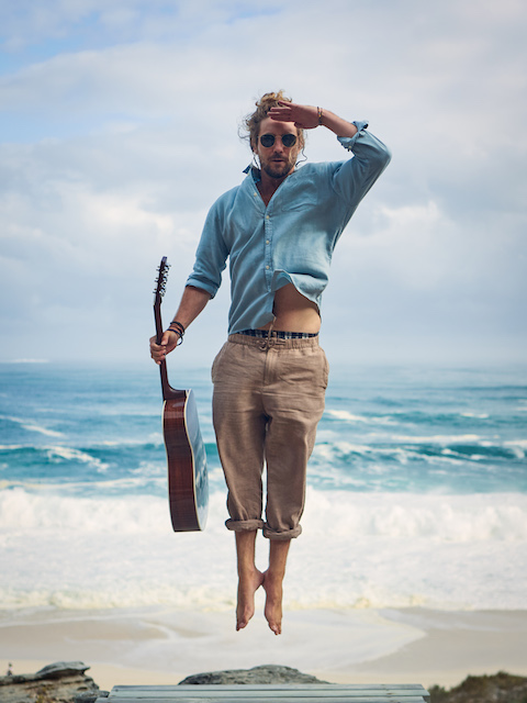 Jeremy Loops wearing a denim shirt jumping and holding a guitar