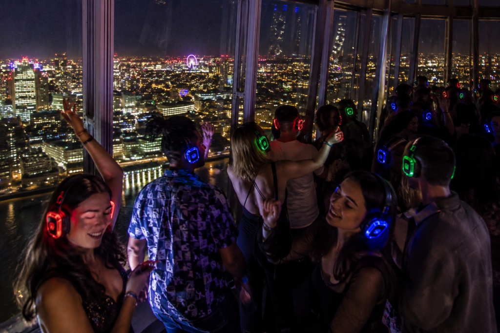 Happy people at a silent disco inside The Shard at night, wearing glowing red, blue and green headphones with panoramic views of London through the glass windows.