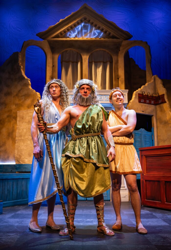 Three actors portraying Zeus, Hebe and Greg in the comedy theatre production Oh Zeus!, set against an Ancient Greek-inspired stage.